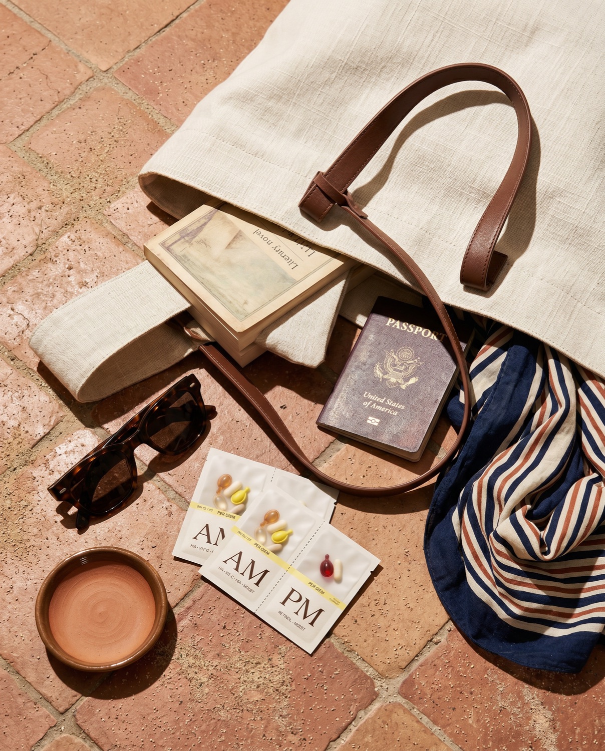 Cream linen tote tipped open on terracotta tile with a passport, three Layover sachets, sunglasses, paperback, and a striped scarf.