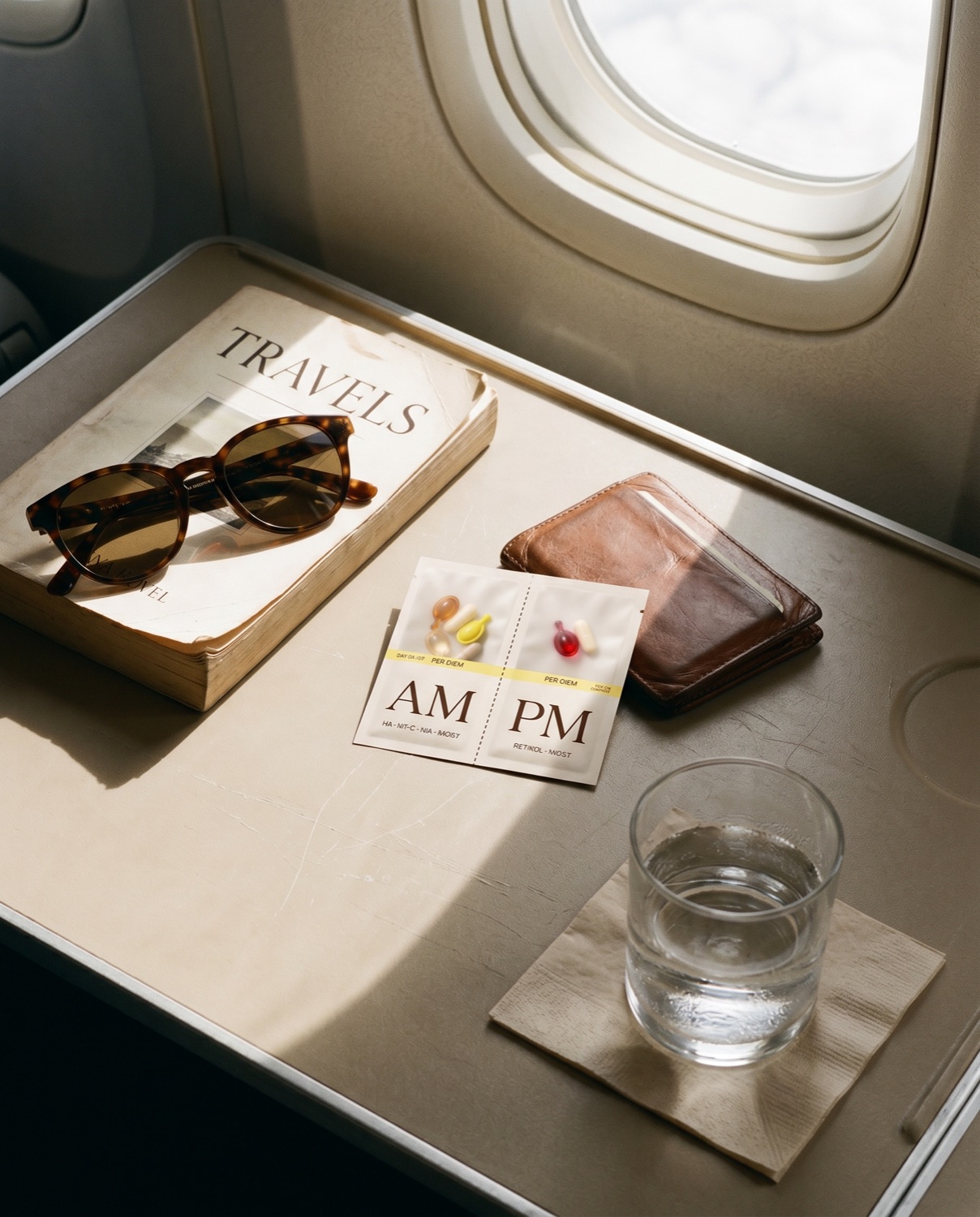 Aircraft tray table with a Travels paperback, tortoiseshell sunglasses, leather card wallet, a Layover sachet, and a glass of water in hard light from the cabin window.