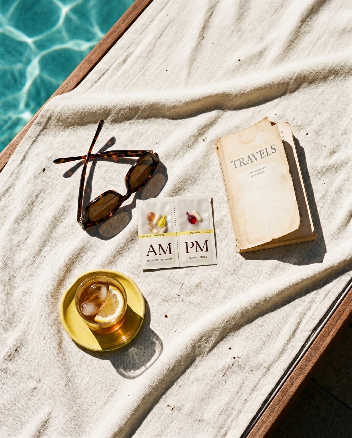Poolside linen towel with tortoiseshell sunglasses, a paperback titled Travels, a Layover sachet, and a banana-yellow ceramic dish.